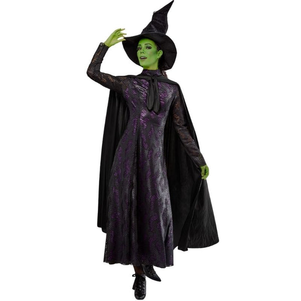 Woman with green skin, wearing the Elphaba Wicked costume, standing in a pose with one hand on the wide-brimmed textured hat. The costume includes an ornate black dress with a lacy texture, purple under-dress, and separate cape, against a white background