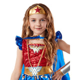 A young girl modeling a Wonder Woman costume against a plain white background. The costume features a red sequined bodice with gold trim, a blue skirt with gold patterns, a blue cape, a gold tiara with a red star, and a silver metallic arm piece.